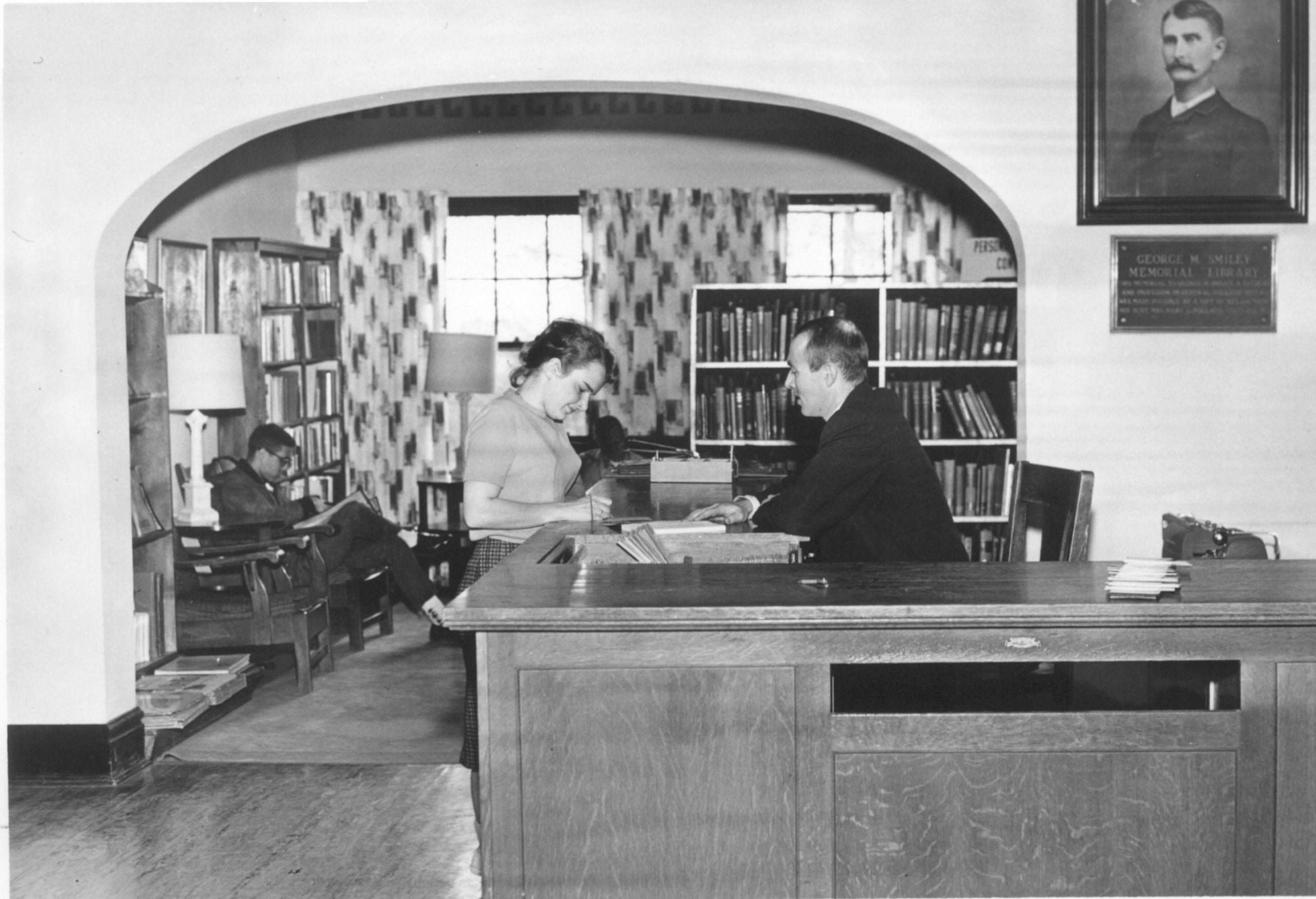 Black-and-white photograph of a male librarian assisting a female student at the circulation desk