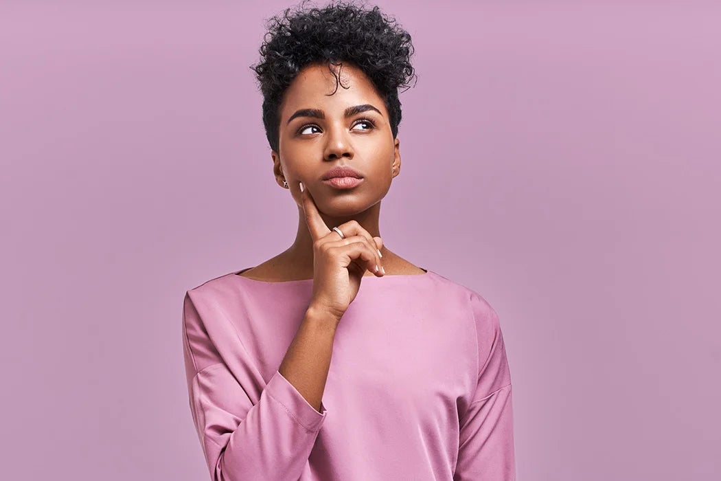 A thoughtful young woman in a pink top stands against a pastel background, touching her chin as if pondering a question. Her expression is reflective, suggesting a moment of contemplation or decision-making.